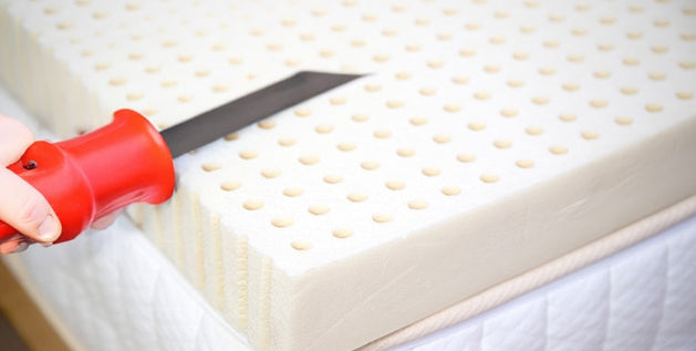 A hand uses a serrated knife to cut a mattress topper to fit the bed, slicing through the latex foam edge.