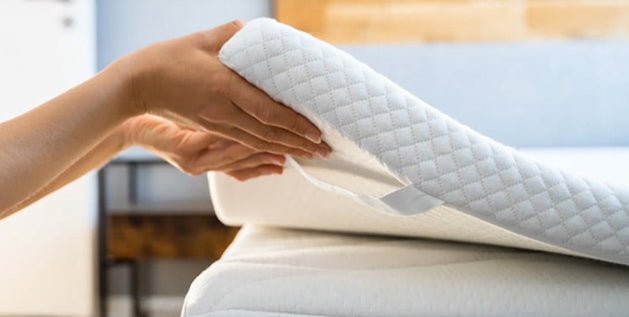 Hands lifting a mattress topper's corner to check its weight and thickness before placing it on the bed.
