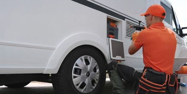 Technician in orange workwear uses a diagnostic laptop while inspecting an RV exterior compartment during dewinterizing.