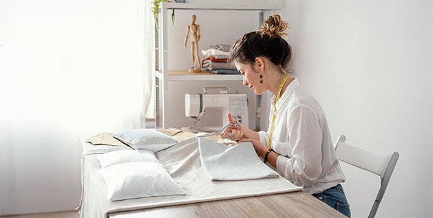 A woman trimming linen fabric for a homemade pillow at a table with sewing tools in the background.