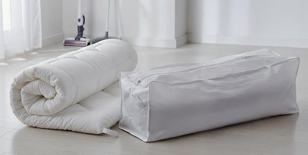 Rolled quilted mattress topper beside a zippered storage bag on a wooden floor.