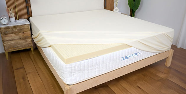 Turmerry mattress topper on a bed, with corner of mattress protector lifted to show the latex foam layer beneath.
