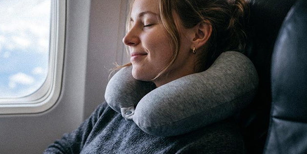A woman sleeping on an airplane with her neck resting on a gray U-shaped travel pillow.
