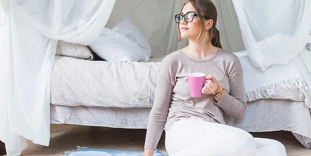 Woman reading a book and holding a coffee mug while relaxing on a blue throw blanket near her bed.