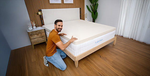 A man kneels beside a bed, fitting a mattress topper onto a mattress with a wooden frame.