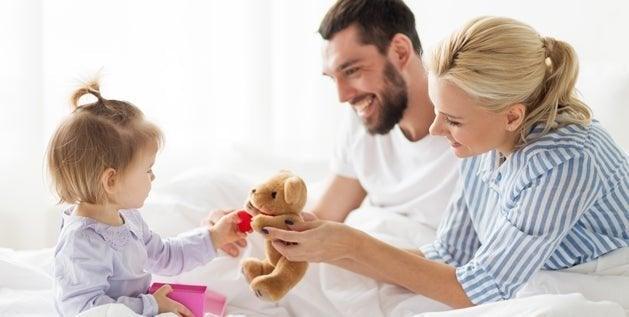 Parents and toddler playing with a teddy bear on white bedding, a cozy sleep setting for a family.