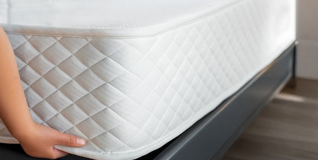 Hand lifting the corner of a white quilted mattress on a dark gray base to check if the mattress is wet underneath.